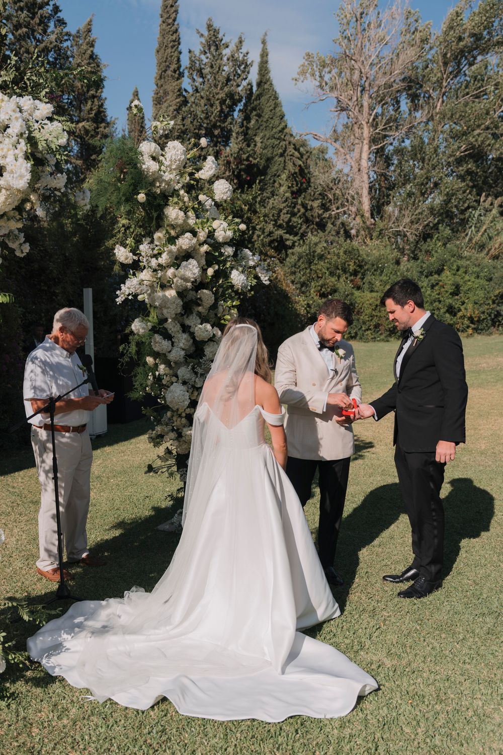An outdoor wedding ceremony under a floral arch with white roses in a garden setting.
