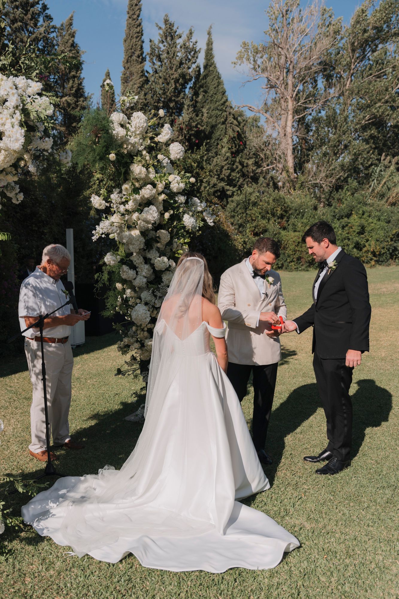 An outdoor wedding ceremony under a floral arch with white roses in a garden setting.
