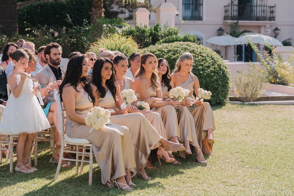 Wedding party members in neutral dresses seated on wooden chairs on a manicured lawn.