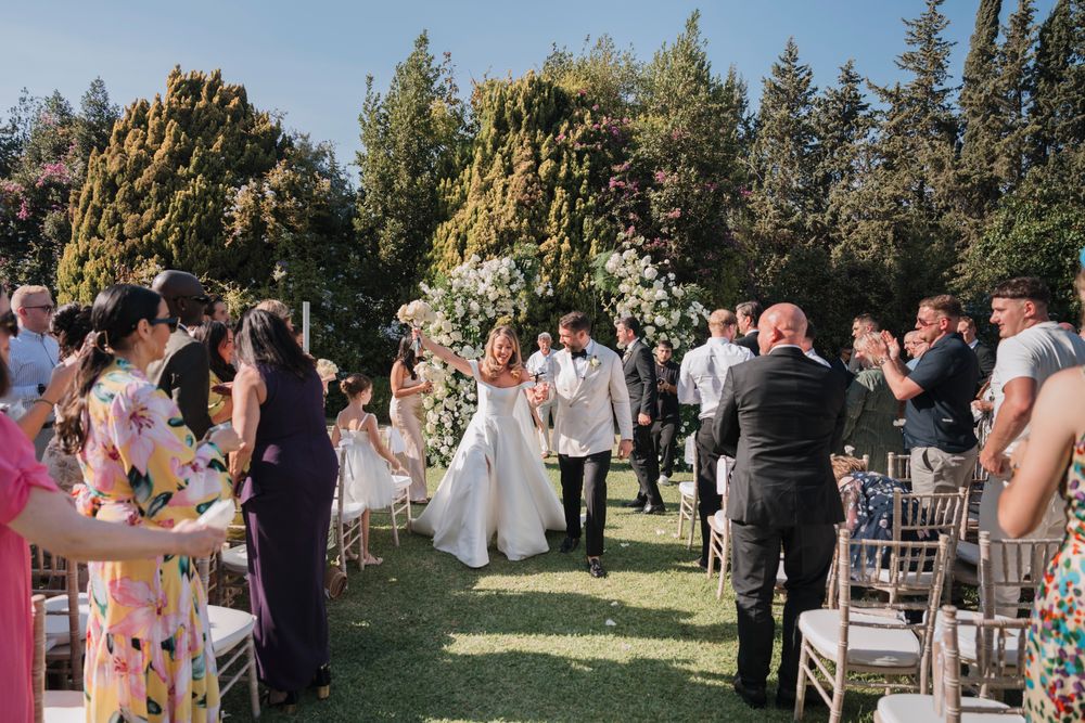 Guests gather for an outdoor garden wedding ceremony under a floral arch on a sunny day.