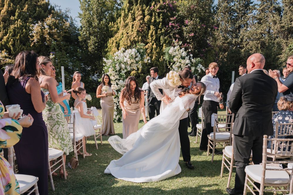 Wedding ceremony taking place outdoors on a lawn with white chairs and floral arrangements.