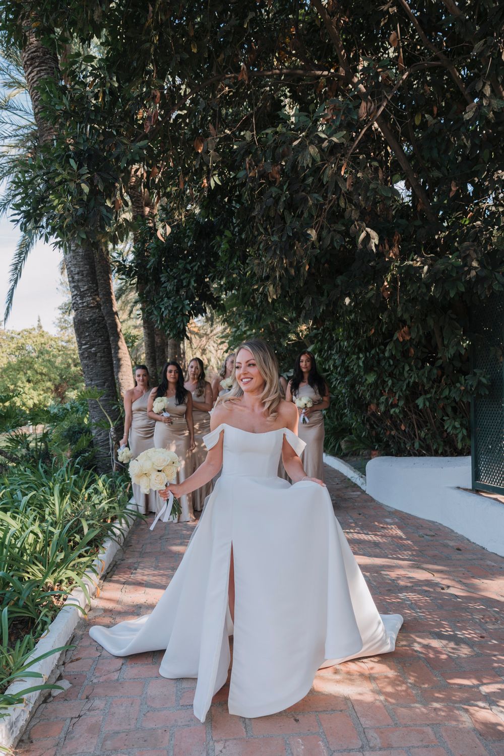 Bride in an elegant white split dress posing on a brick pathway surrounded by lush greenery.