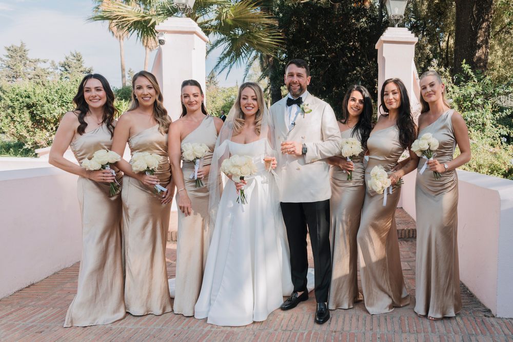 Wedding party poses in champagne-colored dresses against a white Spanish-style building backdrop.