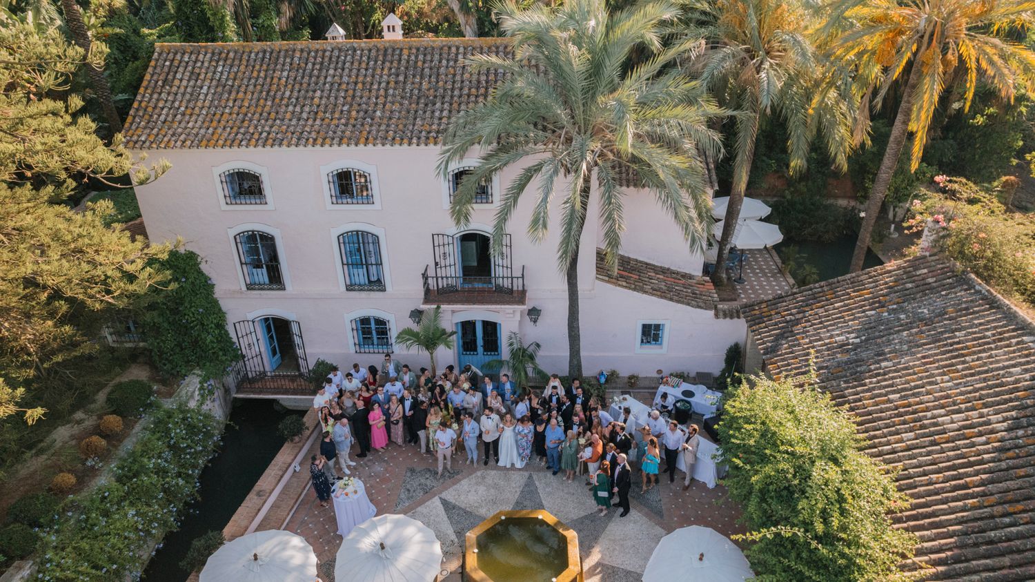 Aerial view of wedding guests gathered outside a white Spanish villa with palm trees.