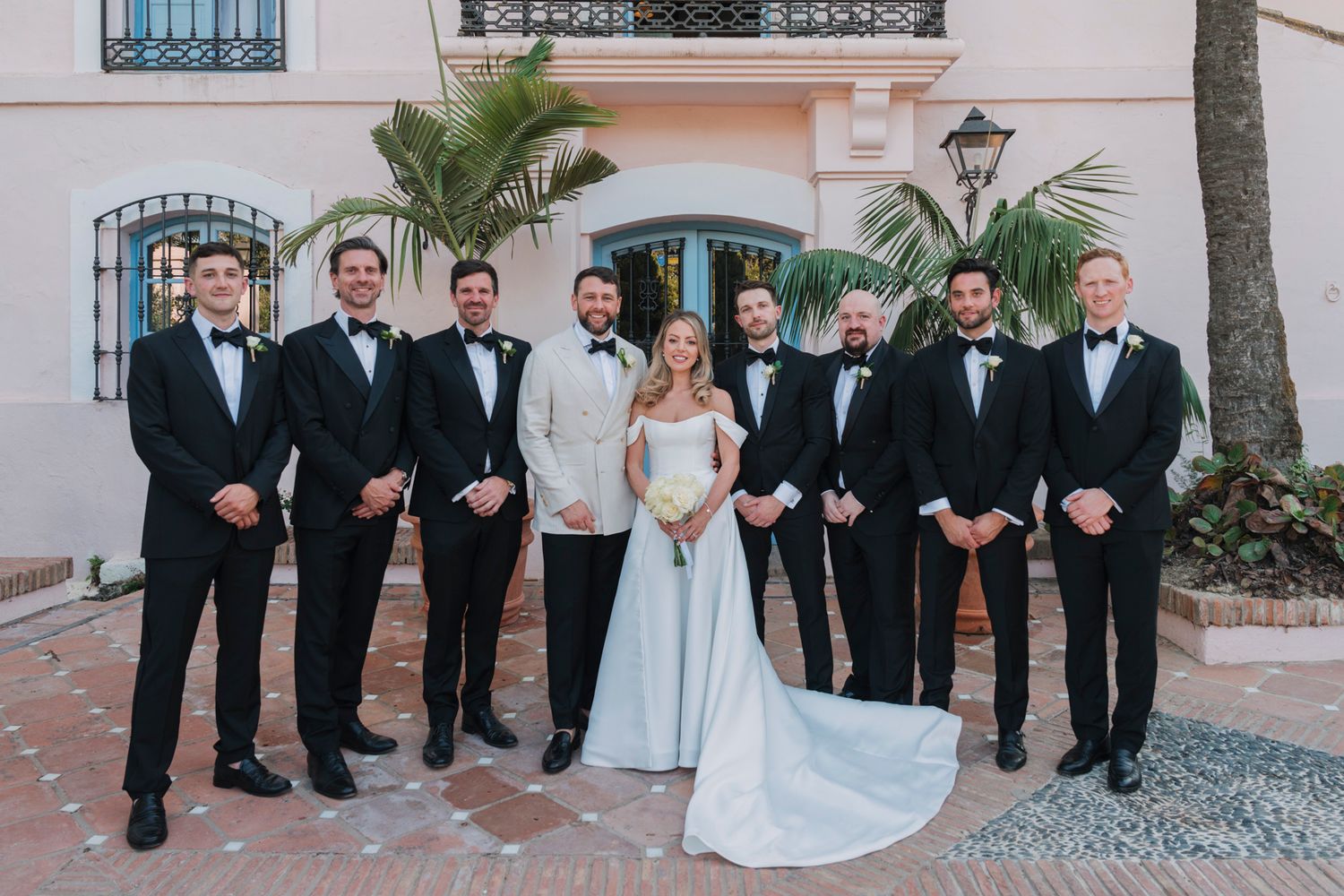 Wedding party in black tuxedos and light blue gown pose together against pink stucco wall with palm trees.