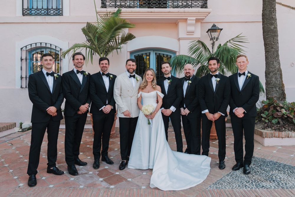 Wedding party in black tuxedos and light blue gown pose together against pink stucco wall with palm trees.
