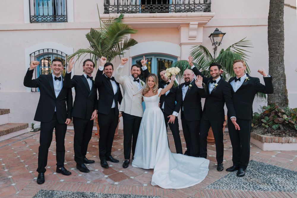 Wedding party jumps joyfully for fun photo outside pink Mediterranean building.