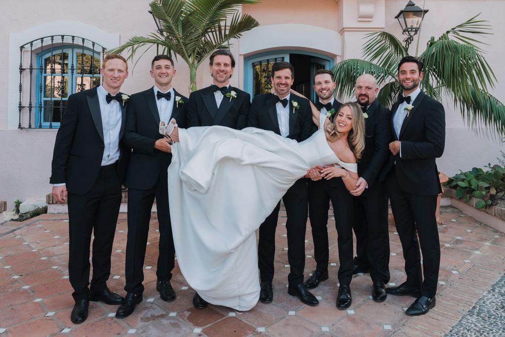 Groomsmen in black tuxedos playfully lift bride in flowing blue dress.