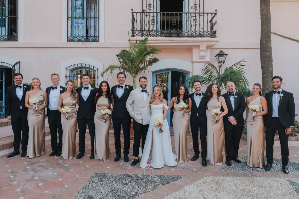 Full wedding party in formal attire poses outside Mediterranean-style building with palm trees.