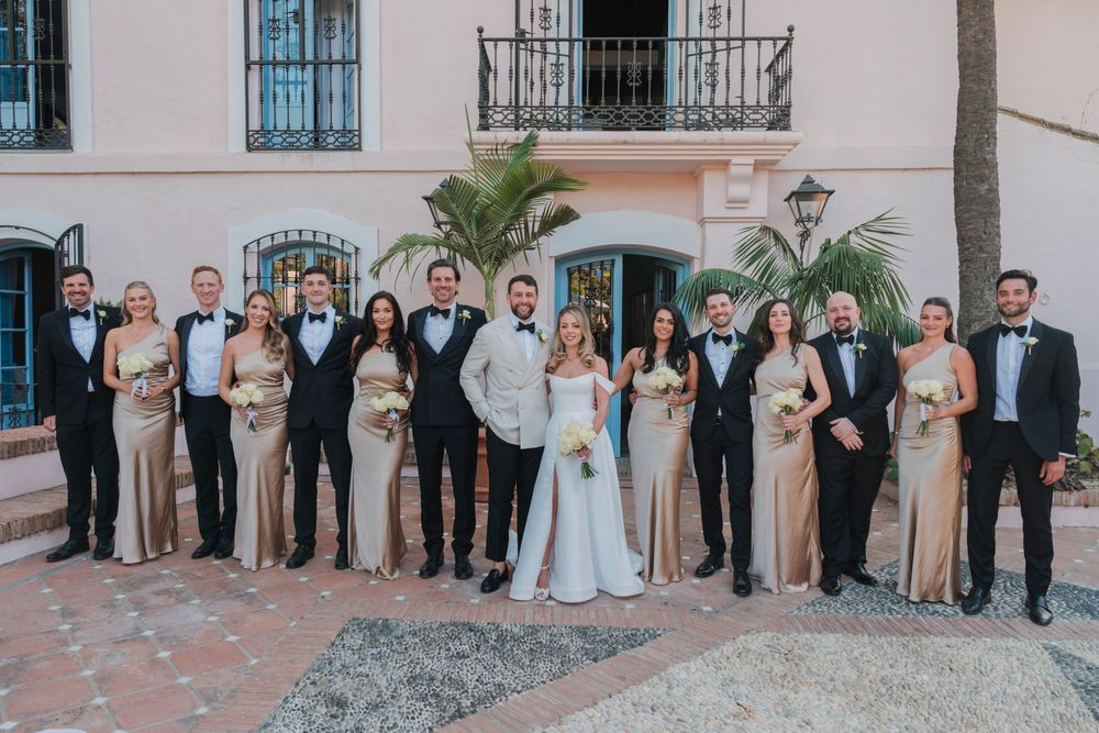 Large wedding party in black tuxedos and champagne dresses stands together against pink building.
