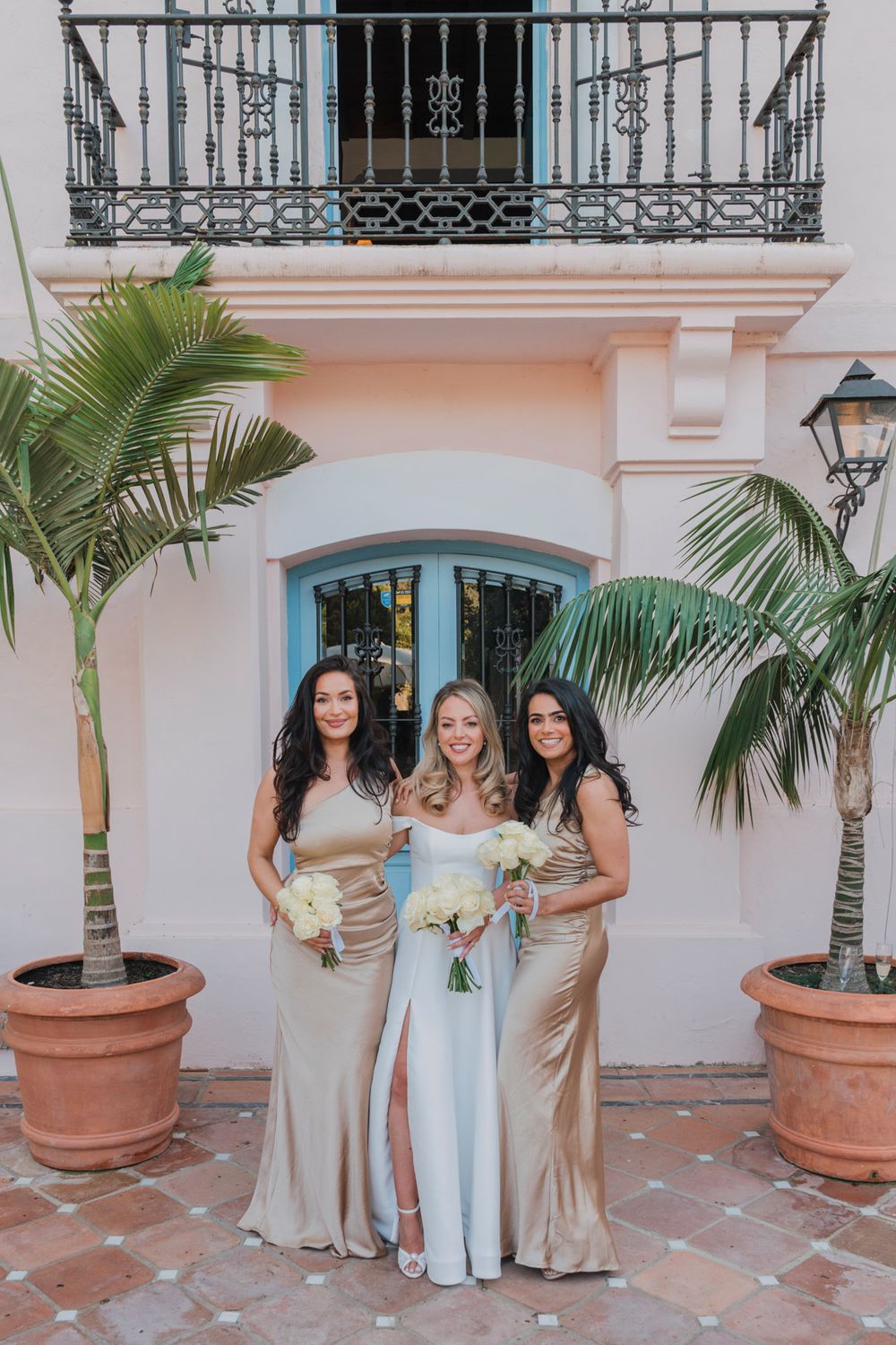 Three women in formal dresses pose between potted palm trees against pink stucco wall.