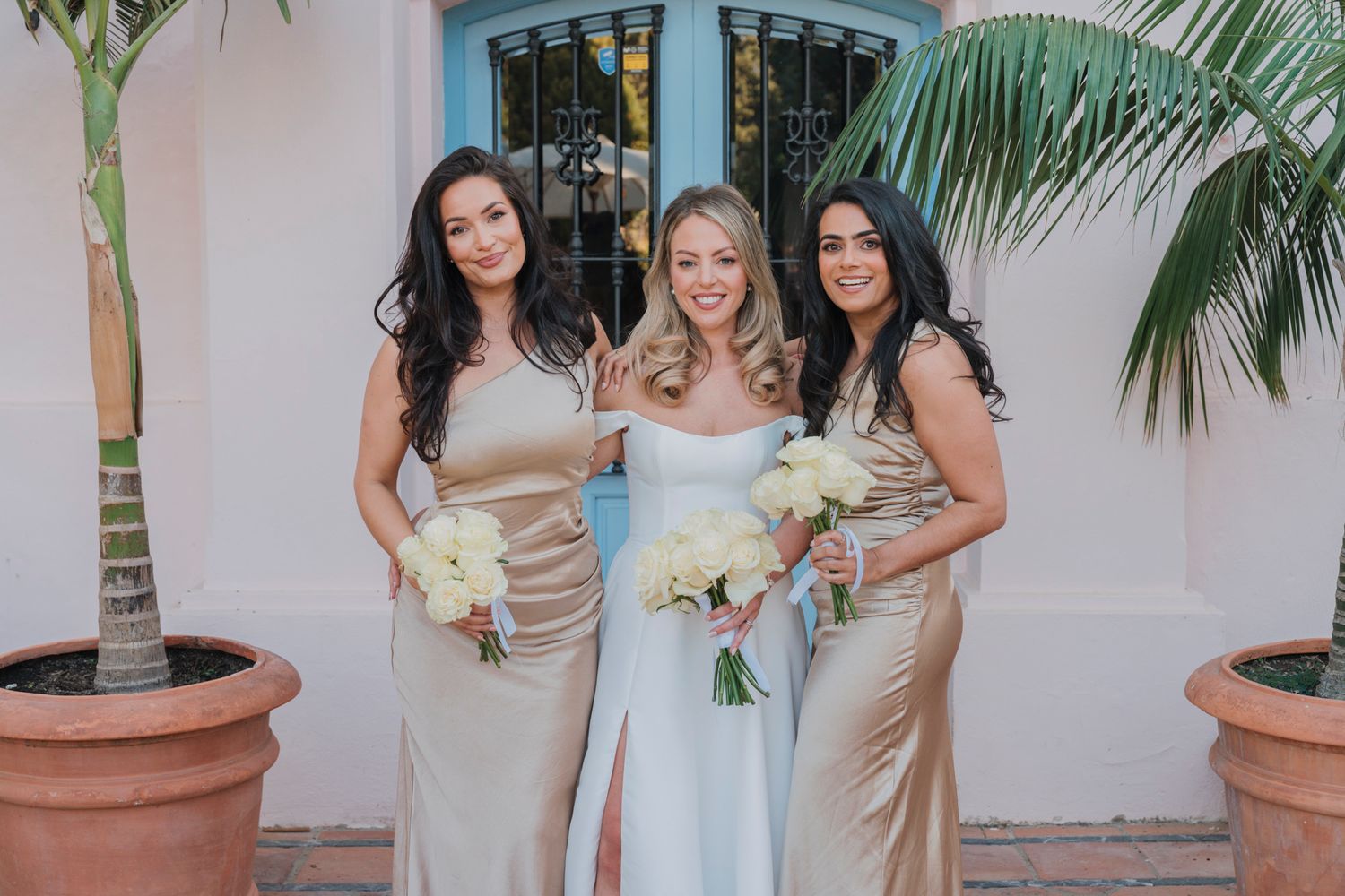 Three women in formal attire hold bouquets while posing against pink Mediterranean building.