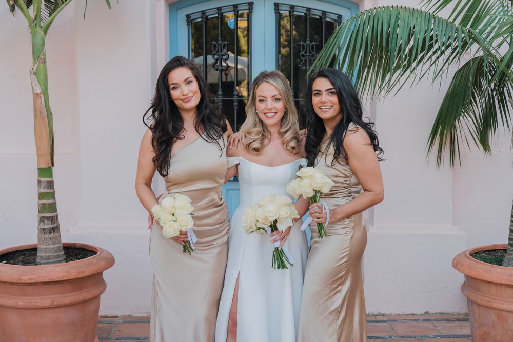 Three women in formal attire hold bouquets while posing against pink Mediterranean building.