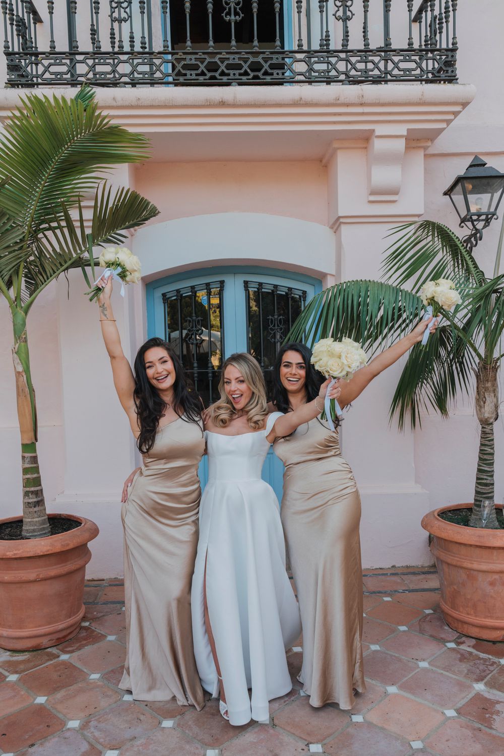 Three women in formal dresses raise bouquets joyfully between palm trees.