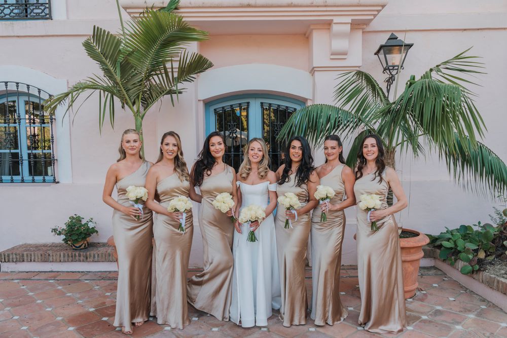 Wedding party in champagne gowns poses with bouquets against pink Mediterranean building.