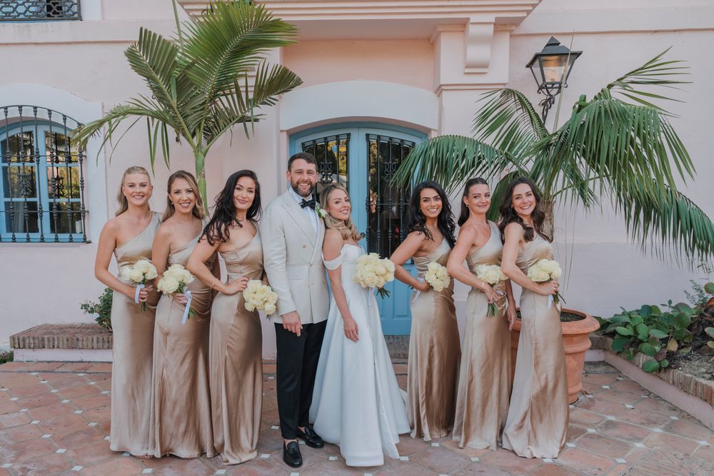 Wedding group in champagne and white dresses poses outdoors against pink stucco wall.