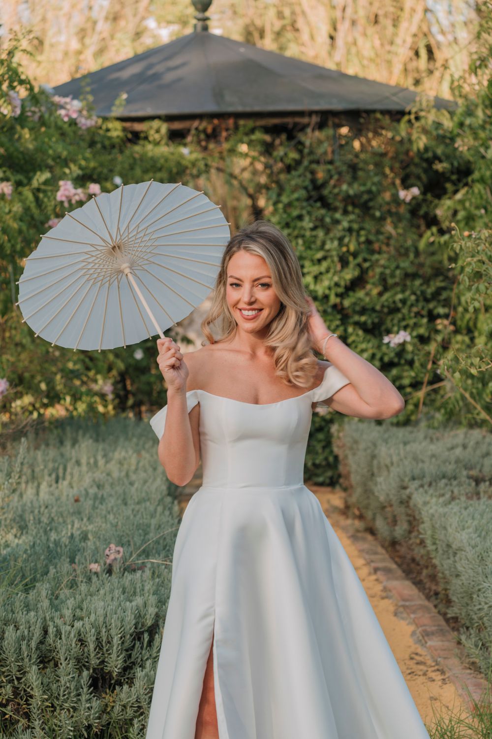 An elegant white wedding dress with a parasol in a garden setting at golden hour.