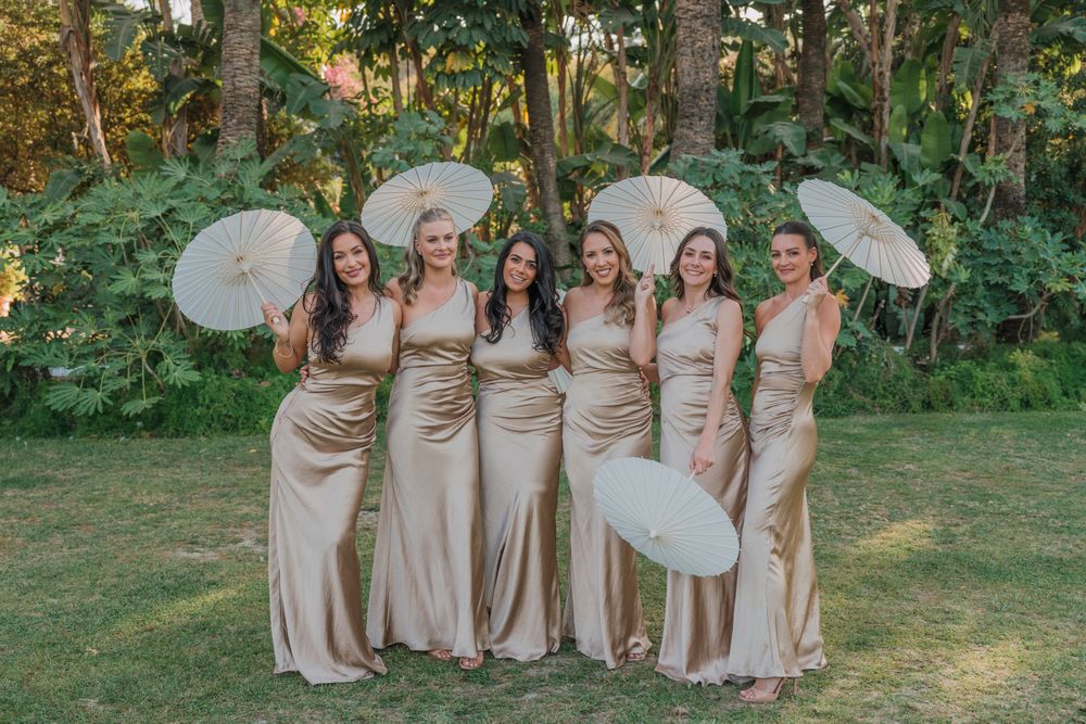 Group photo of bridesmaids in champagne silk dresses holding white parasols in a garden.