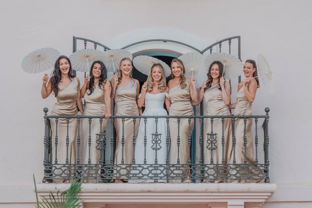Wedding party poses with parasols on an ornate white balcony with decorative railings.