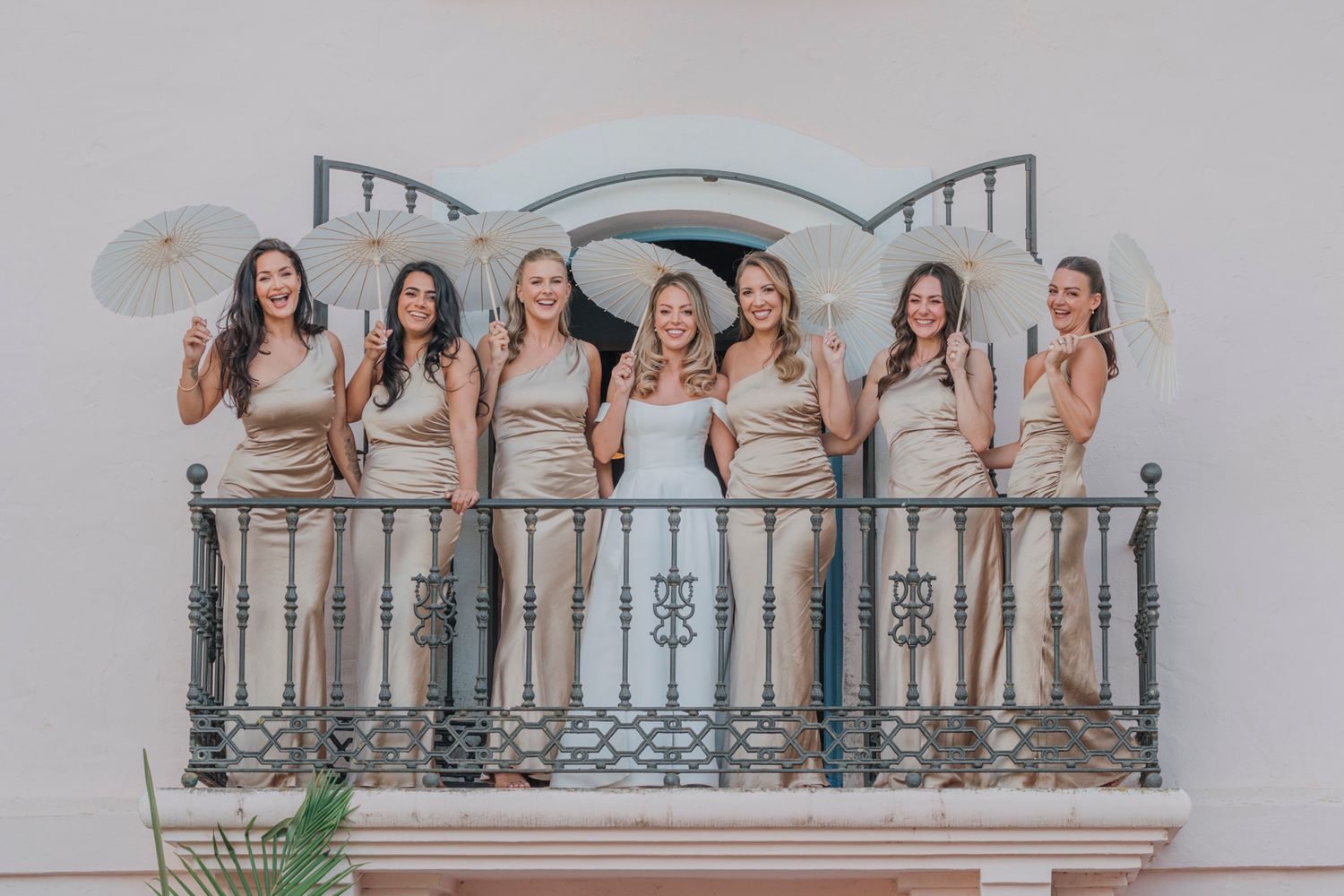 Wedding party poses with parasols on an ornate white balcony with decorative railings.