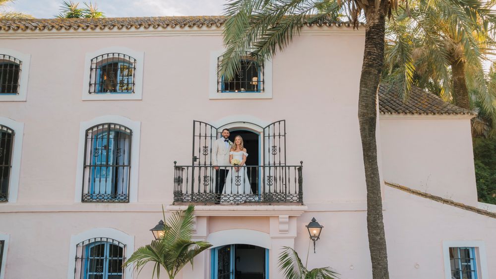 Wedding couple standing on a pink Mediterranean villa balcony with palm trees.