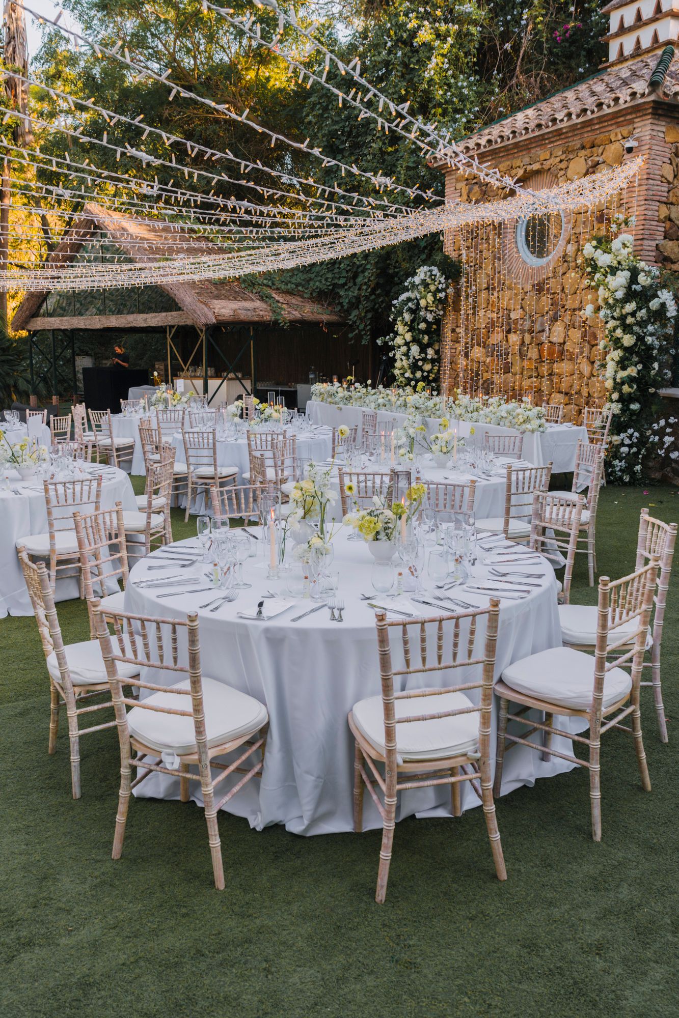 Outdoor wedding reception setup with white linens, string lights, and Chiavari chairs under a romantic canopy.
