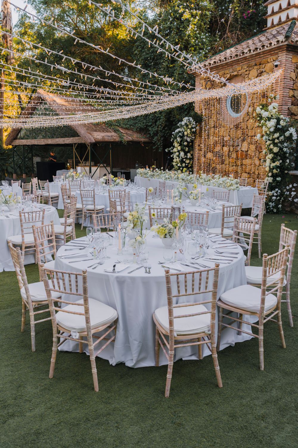 Outdoor wedding reception setup with white linens, string lights, and Chiavari chairs under a romantic canopy.