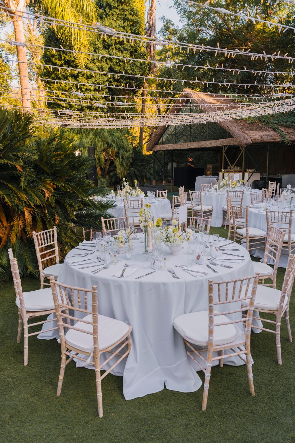 Garden wedding reception tables with white tablecloths and light wooden chairs arranged under twinkling lights.
