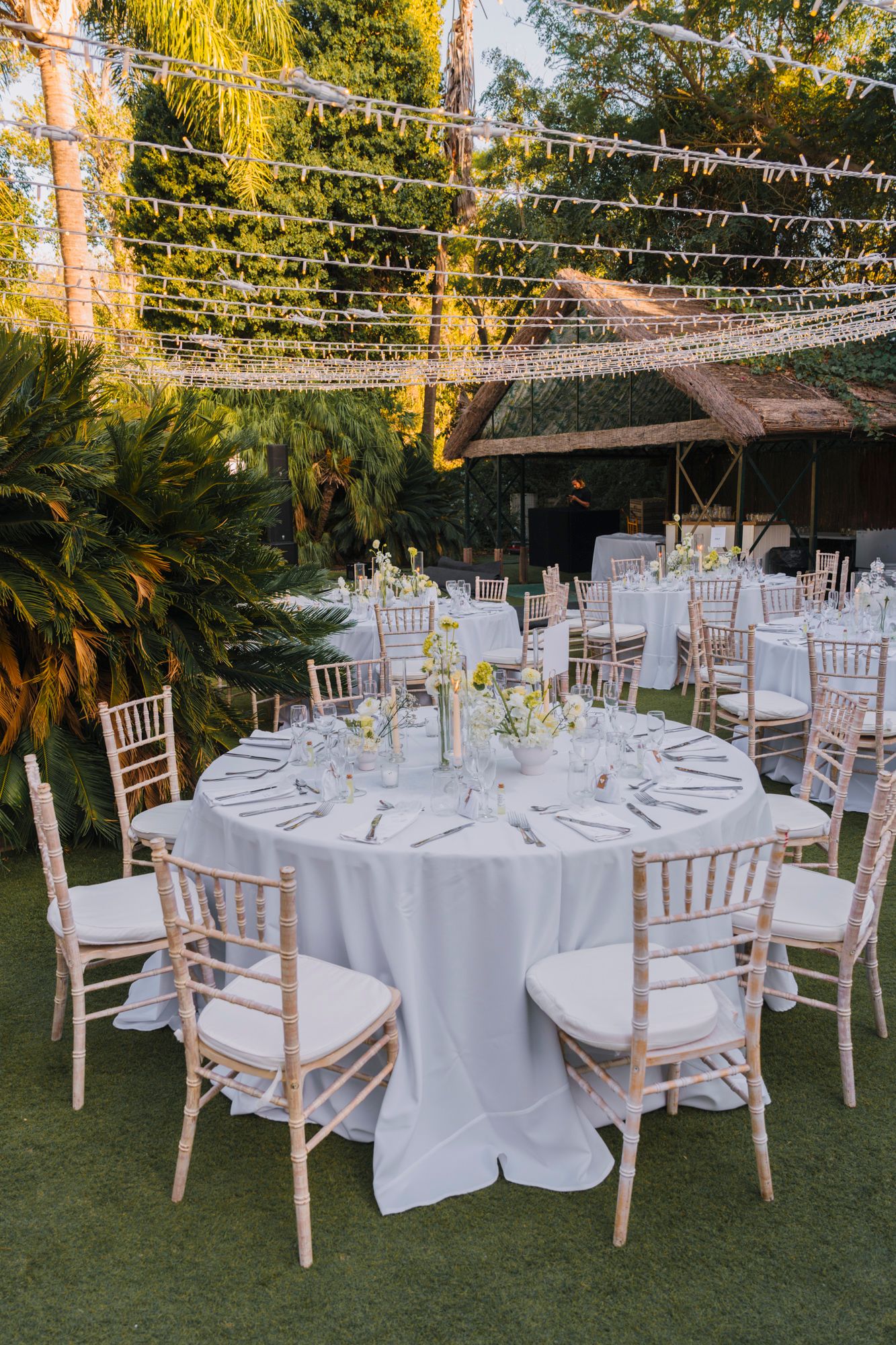 Garden wedding reception tables with white tablecloths and light wooden chairs arranged under twinkling lights.
