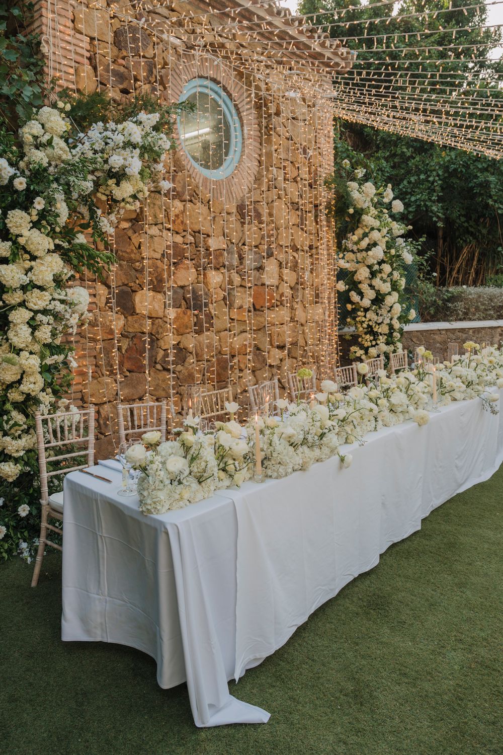 Wedding head table setup against a rustic stone wall with white floral arrangements and string lights overhead.