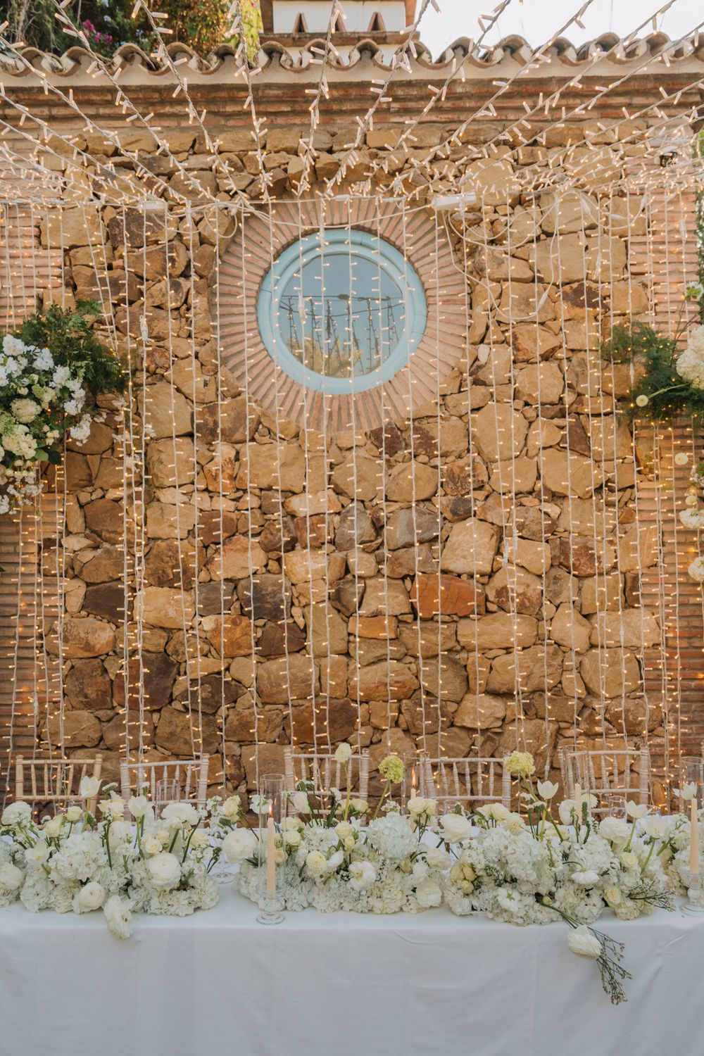 Stone wall backdrop with circular window and cascading white flowers for wedding reception.