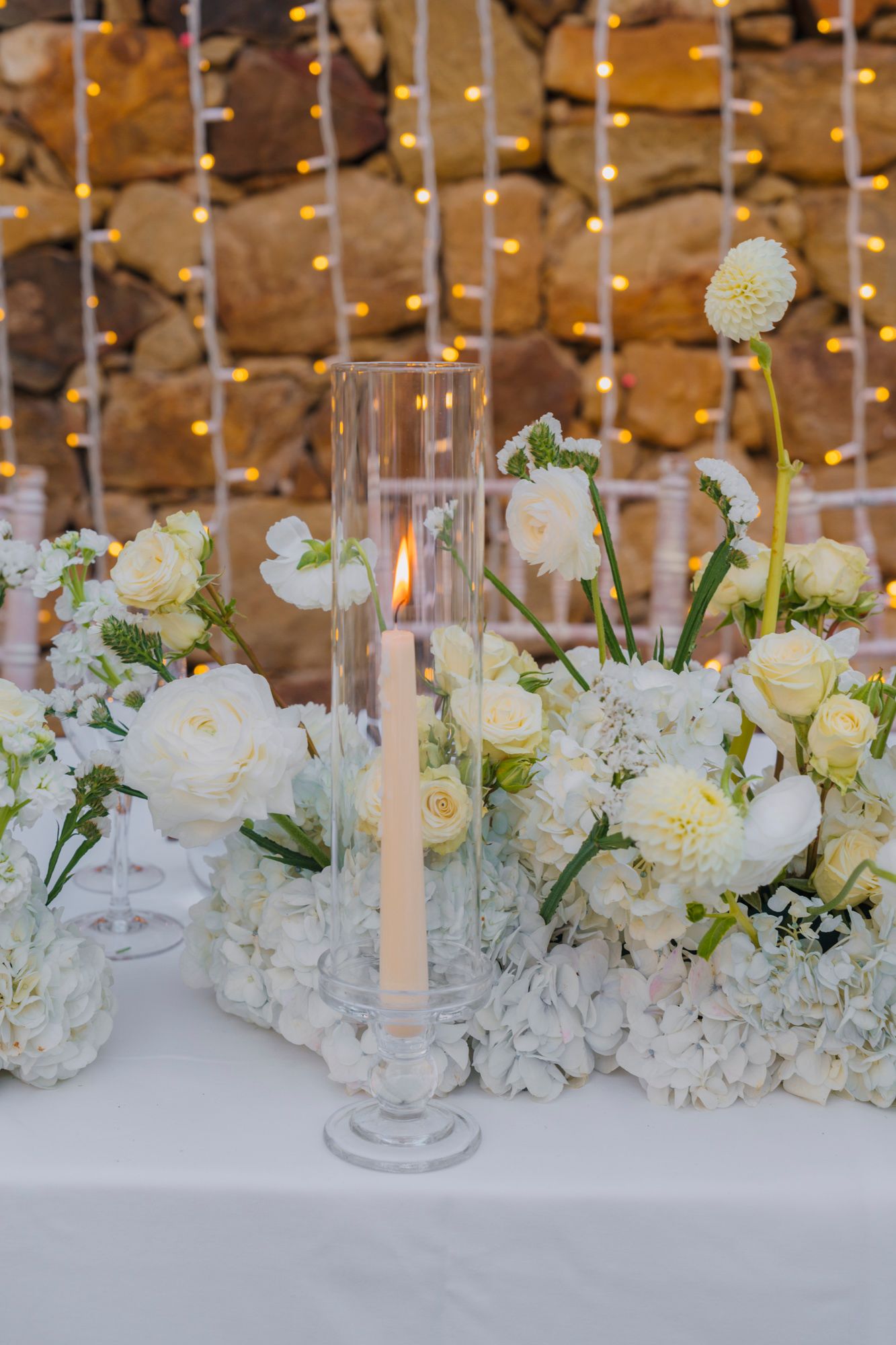 Close-up of elegant white floral centerpiece with glass candleholders and roses on wedding table.
