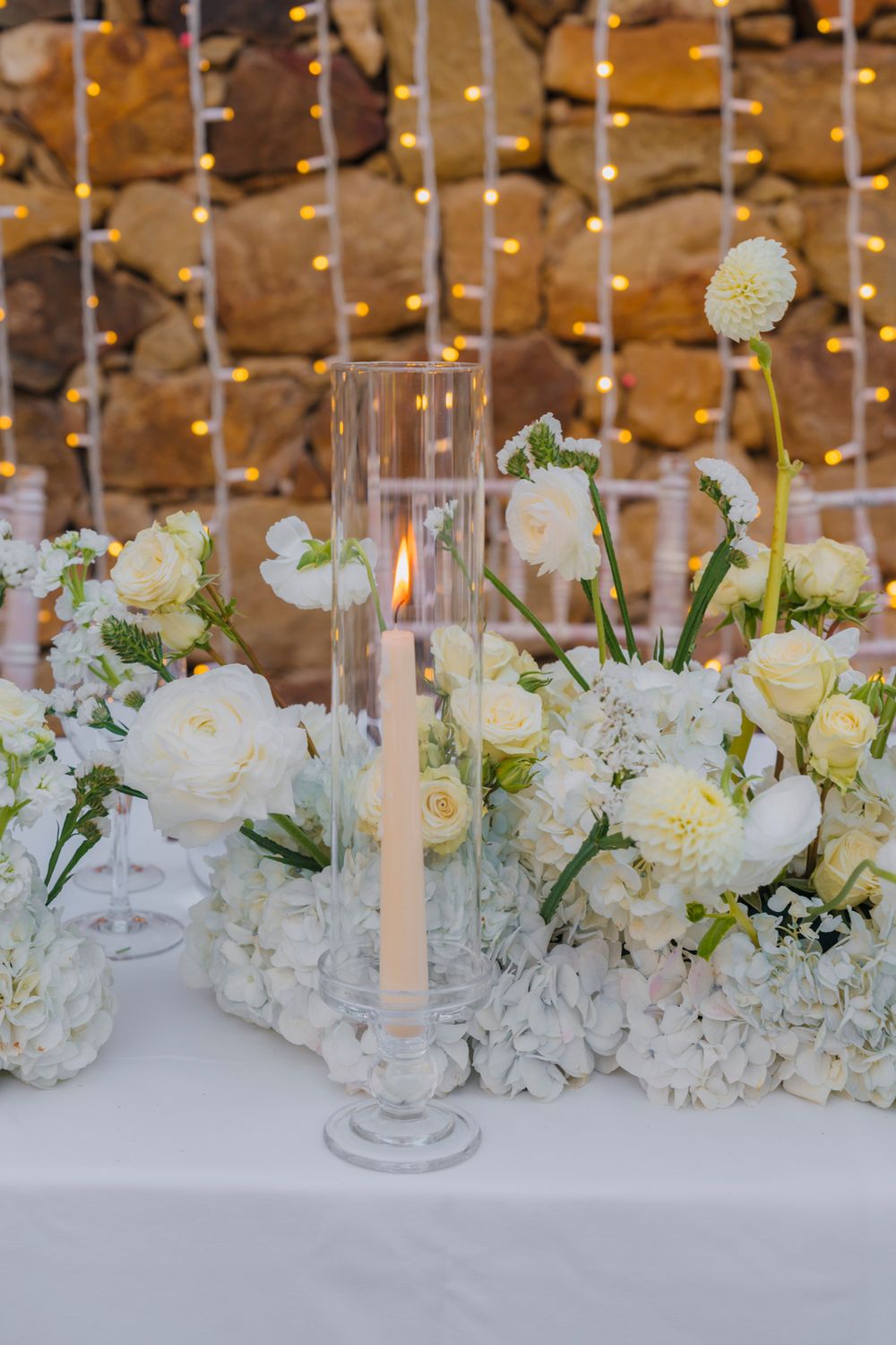 Close-up of elegant white floral centerpiece with glass candleholders and roses on wedding table.