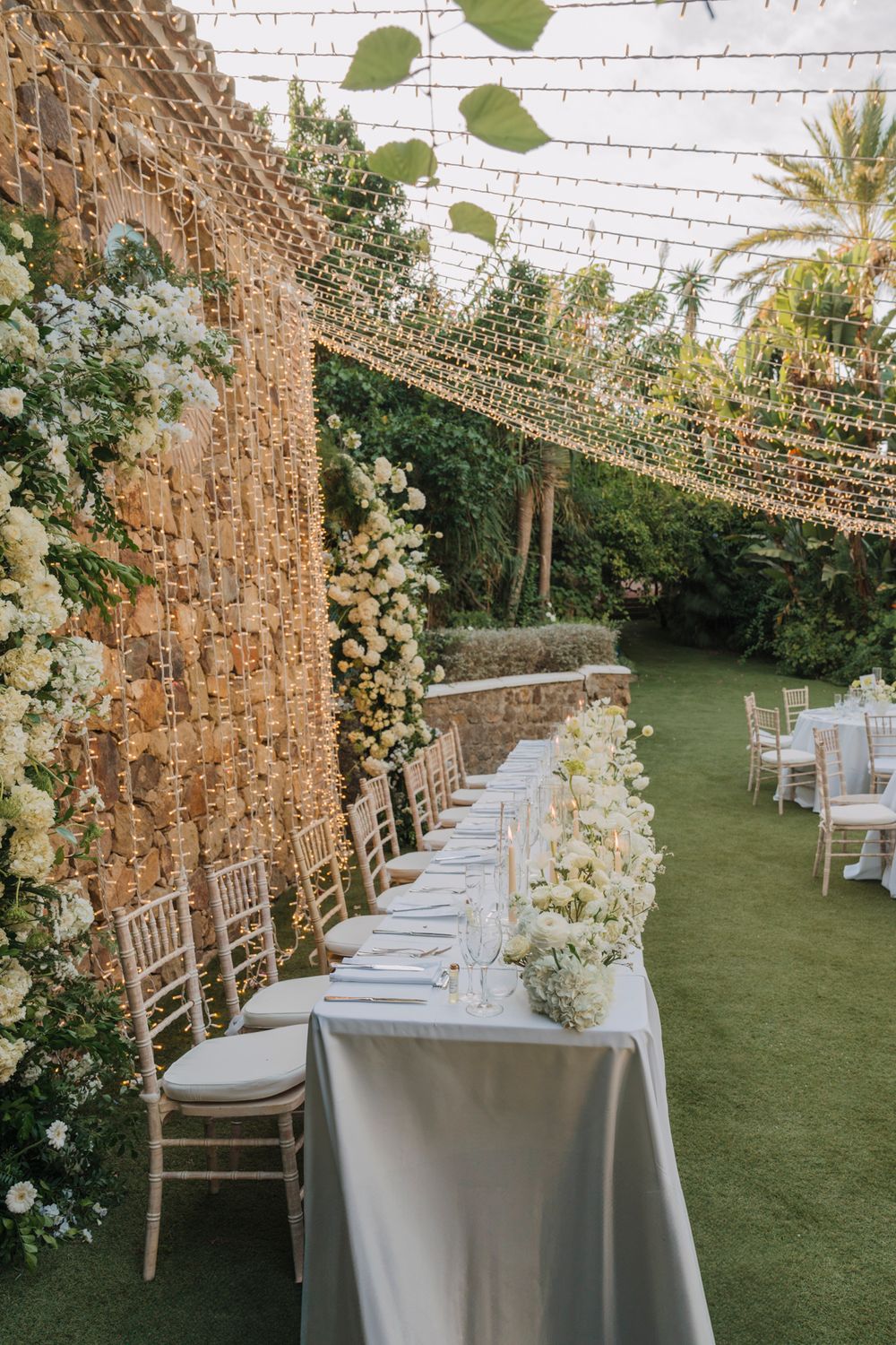 Long wedding table setup along stone wall with string lights and white floral arrangements.
