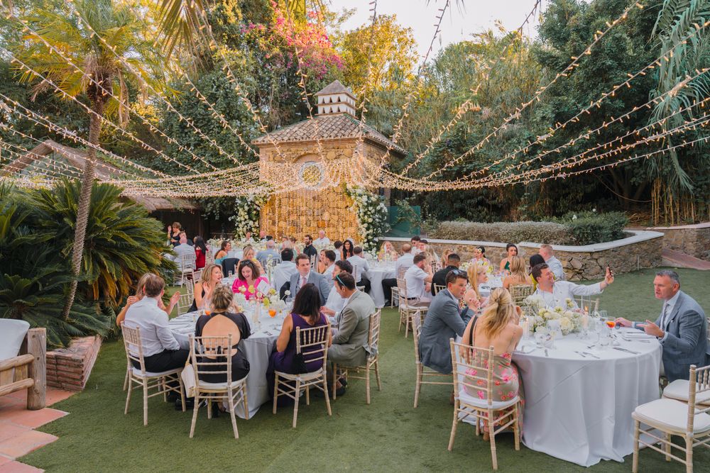 Elegant outdoor wedding reception with string lights and round tables set under a glowing chandelier.