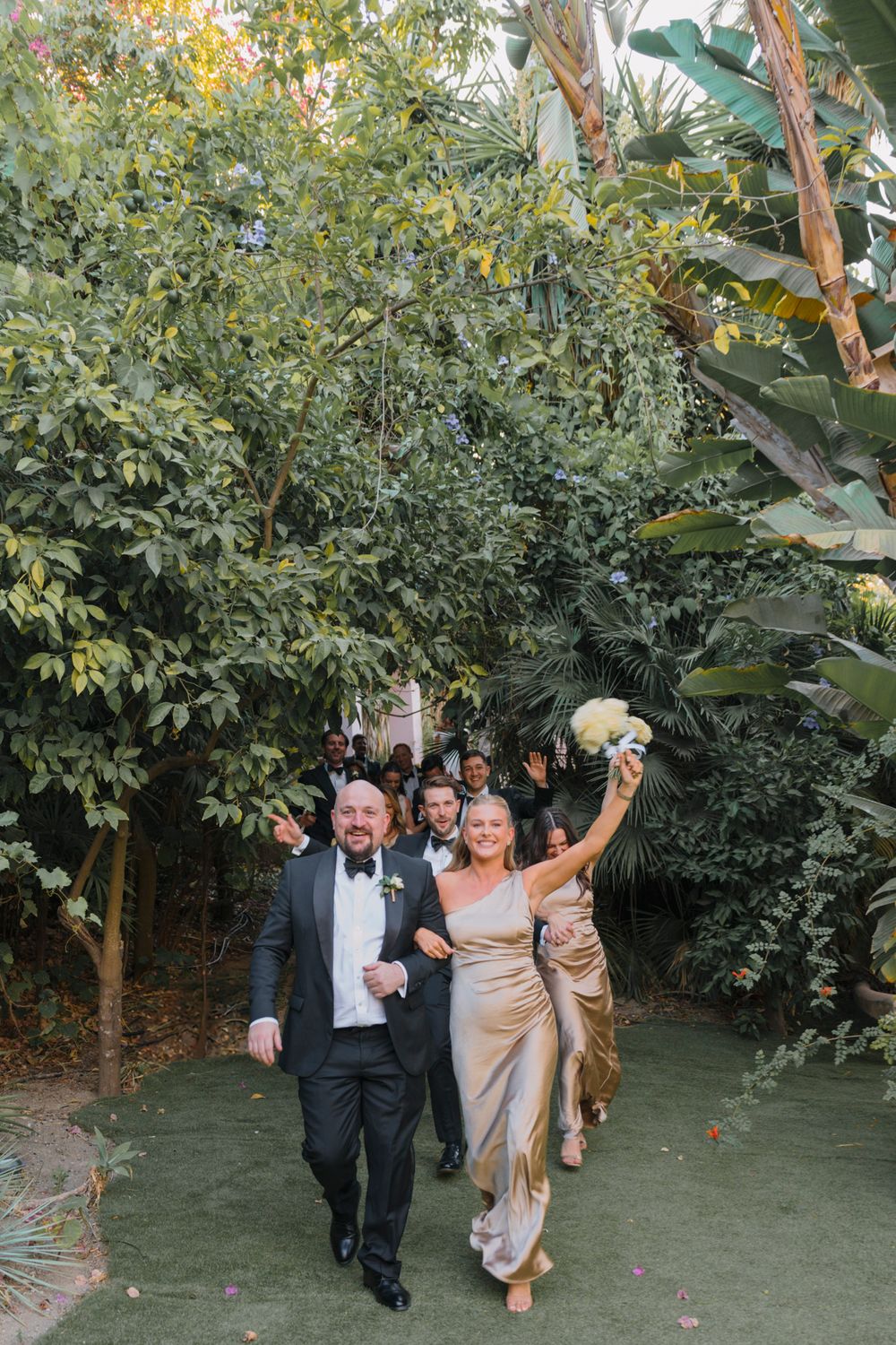 Wedding party walking joyfully through a garden path lined with tropical greenery.