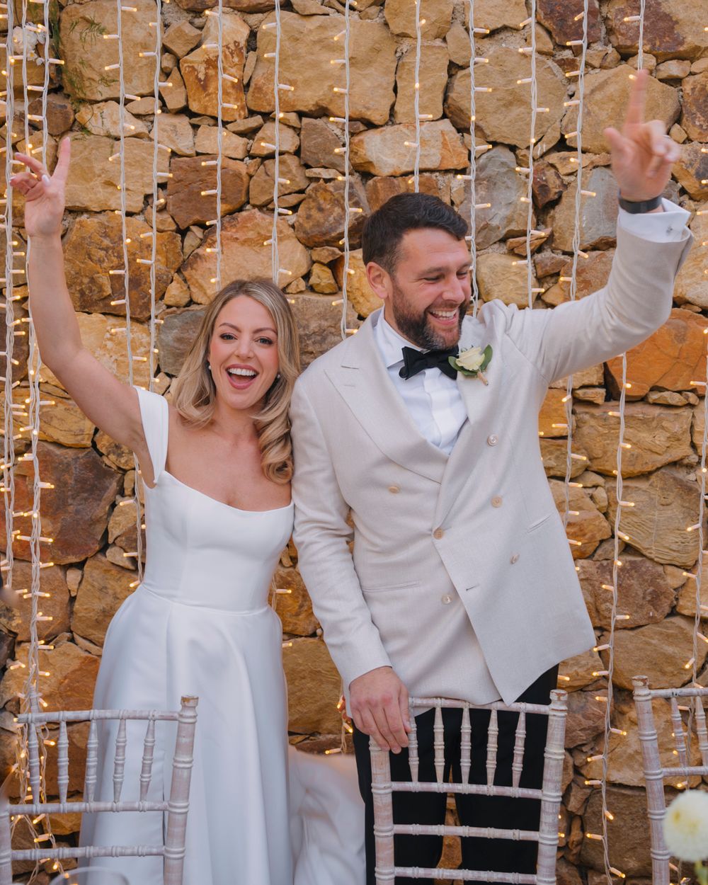A celebratory moment at a wedding reception with string lights and stone wall backdrop.