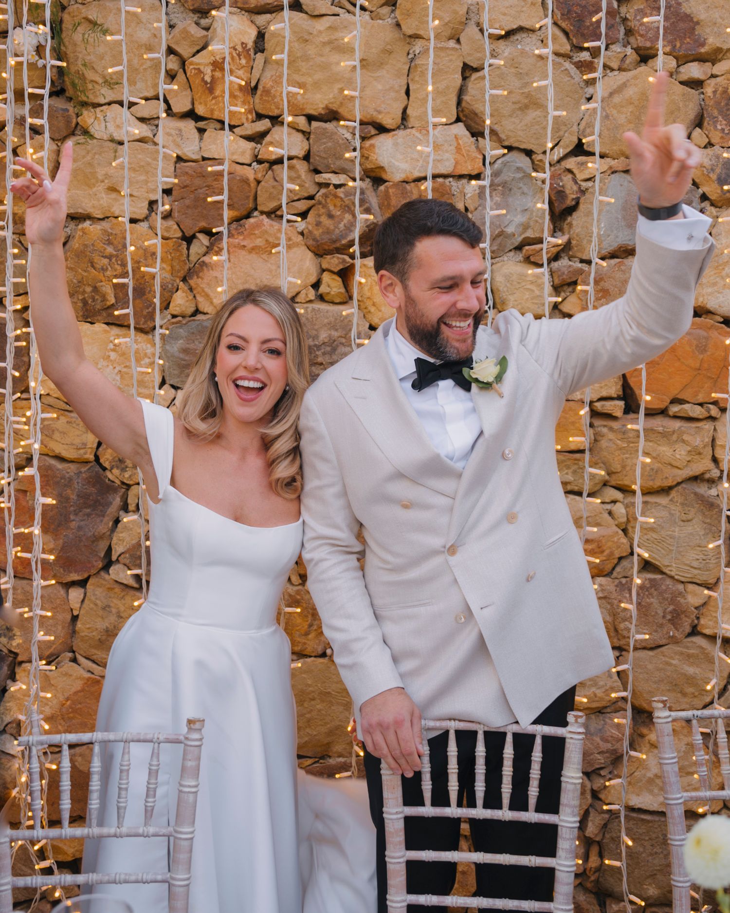 A celebratory moment at a wedding reception with string lights and stone wall backdrop.