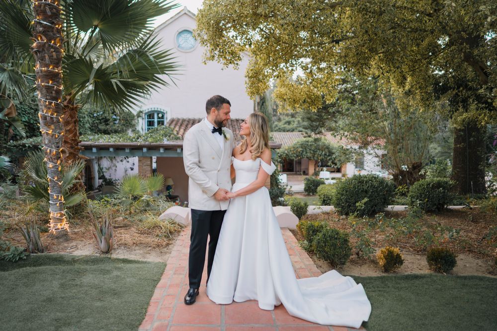 A bride and groom pose together on a brick pathway surrounded by palm trees and lush landscaping.