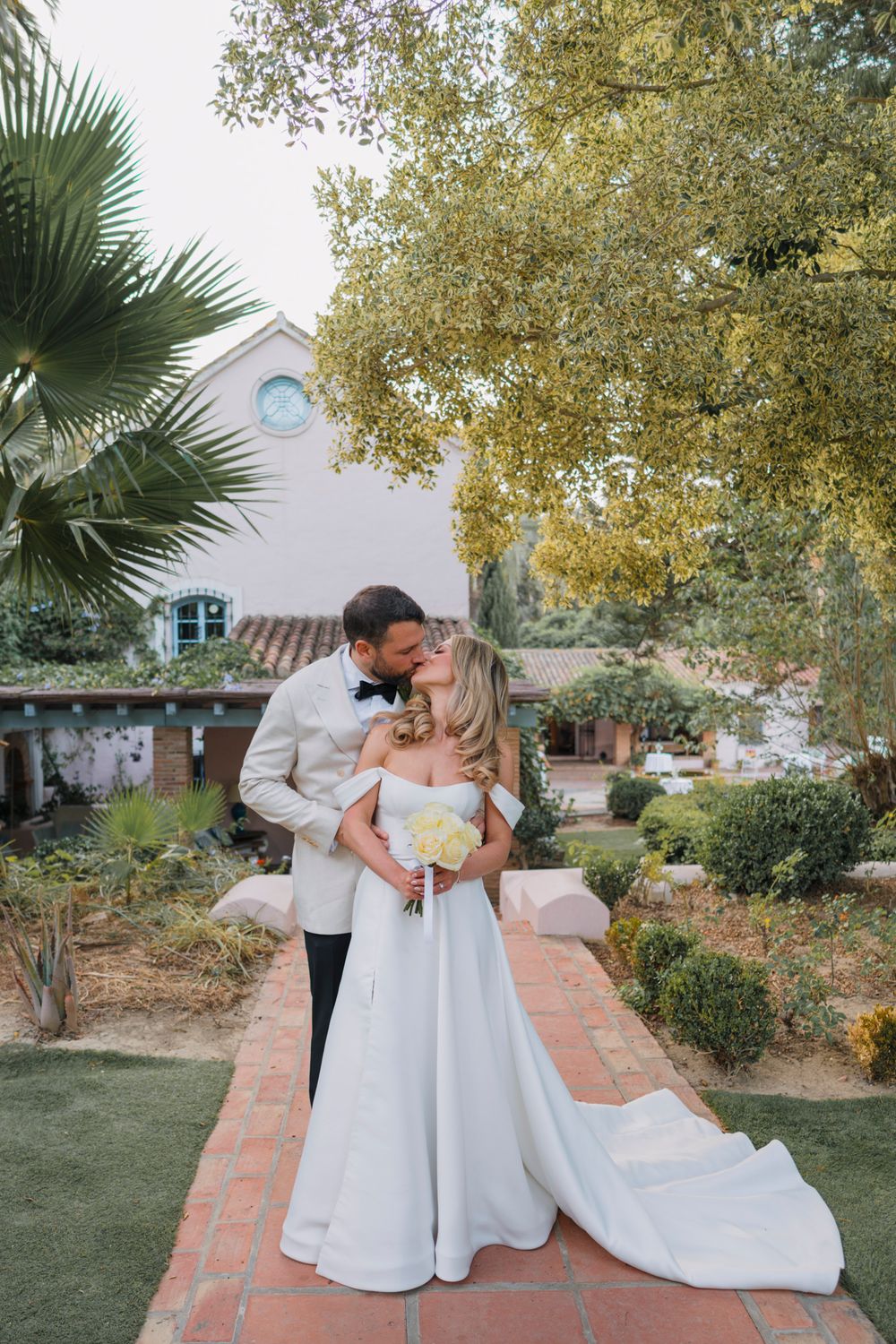 A romantic wedding portrait on a brick path with a Spanish-style building and palm trees in the background.