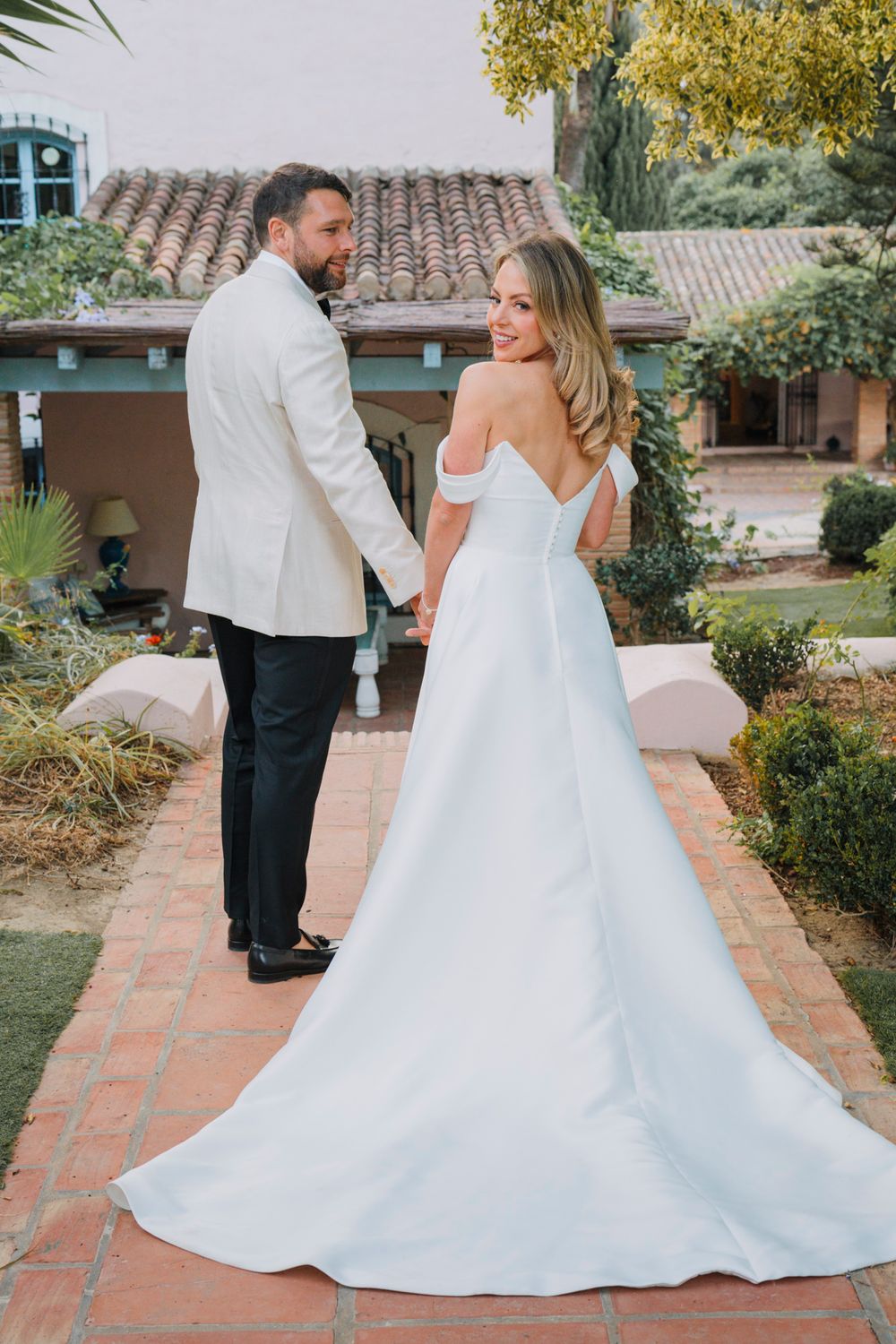 An elegant wedding couple walks hand in hand on a brick pathway with a flowing white gown trailing behind.