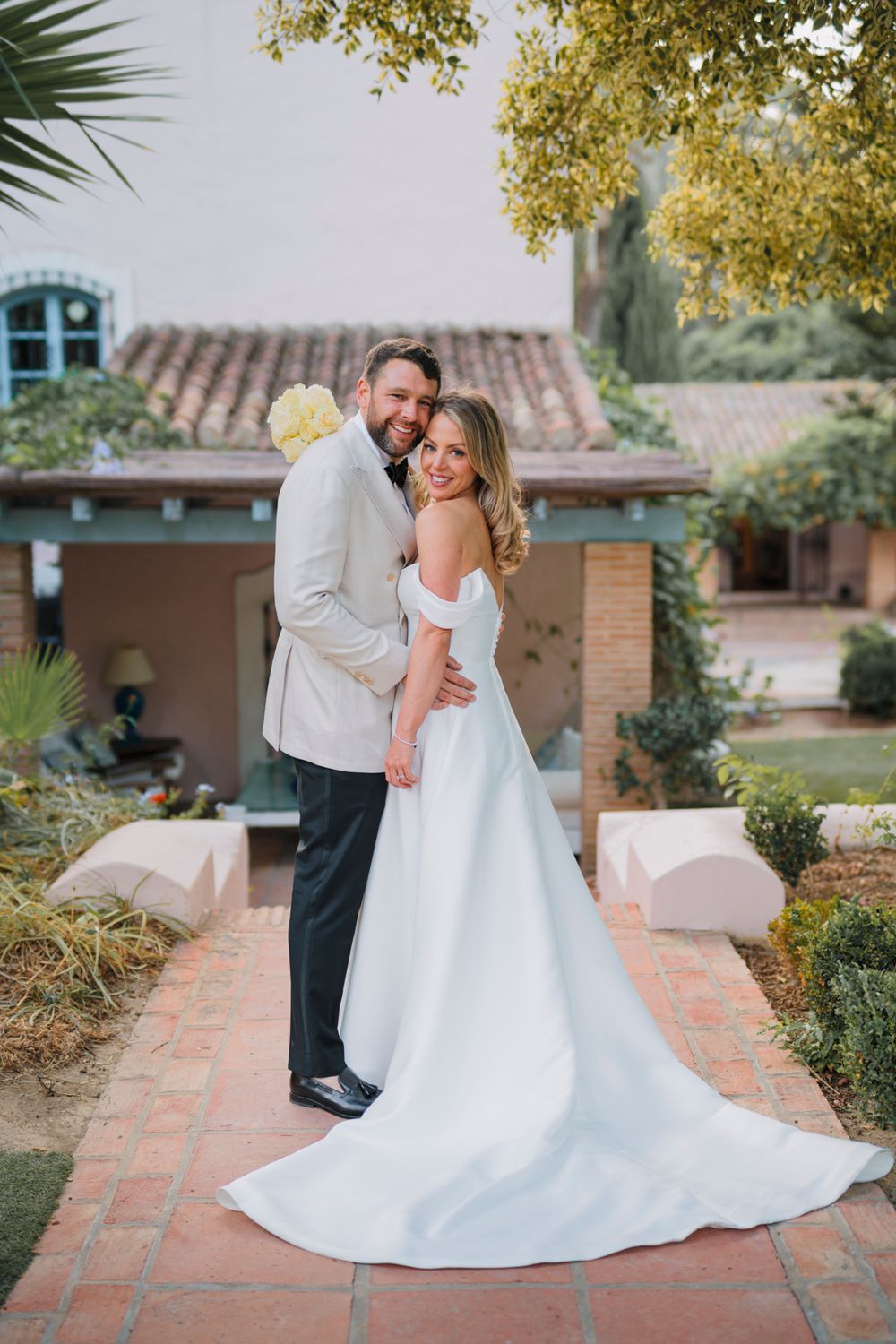 A joyful wedding couple embraces on a brick pathway surrounded by garden landscaping.