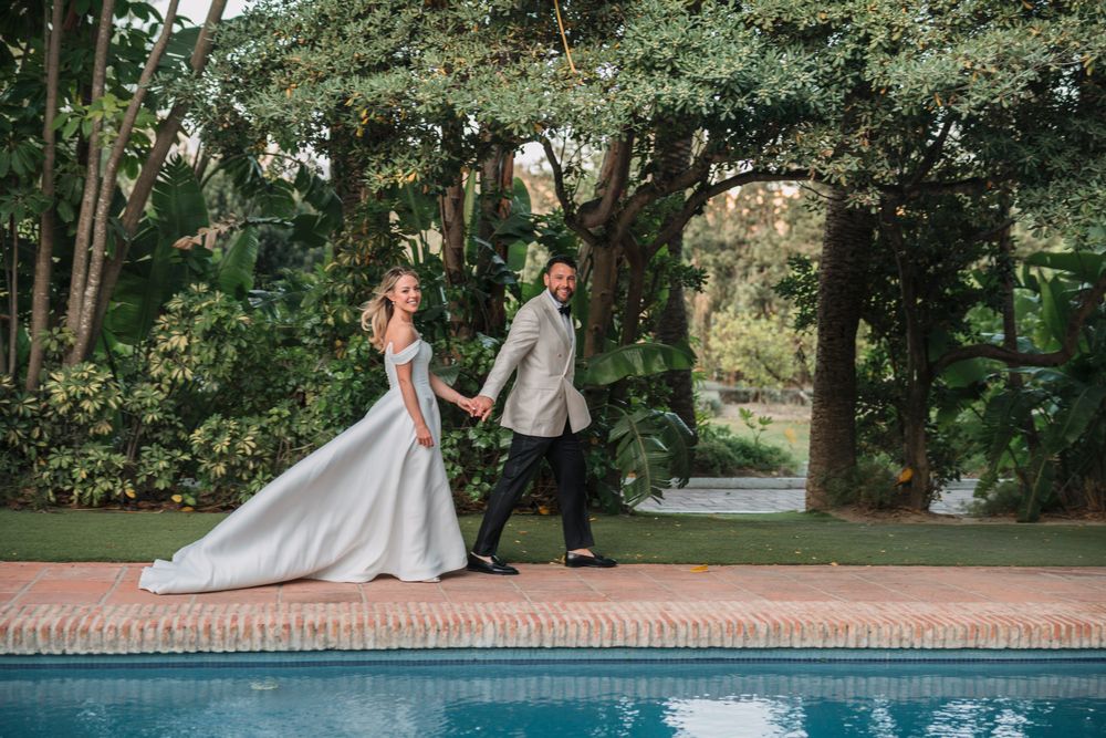 A bride and groom walk along a poolside path surrounded by lush greenery.