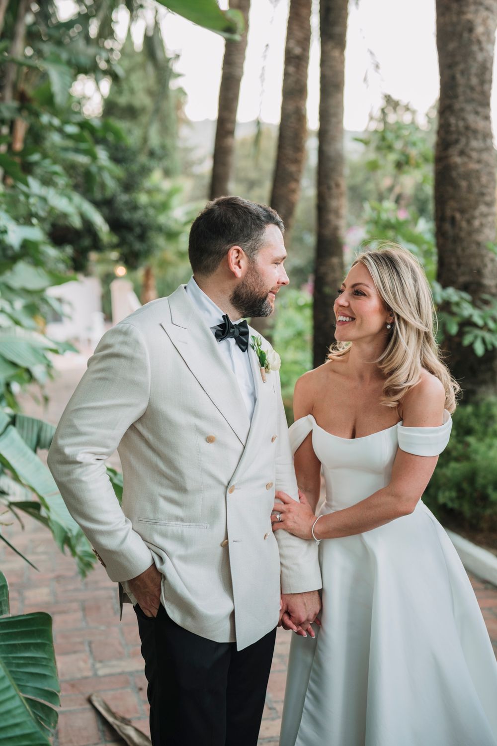 A newlywed couple shares a joyful moment together on a brick path surrounded by lush tropical foliage.