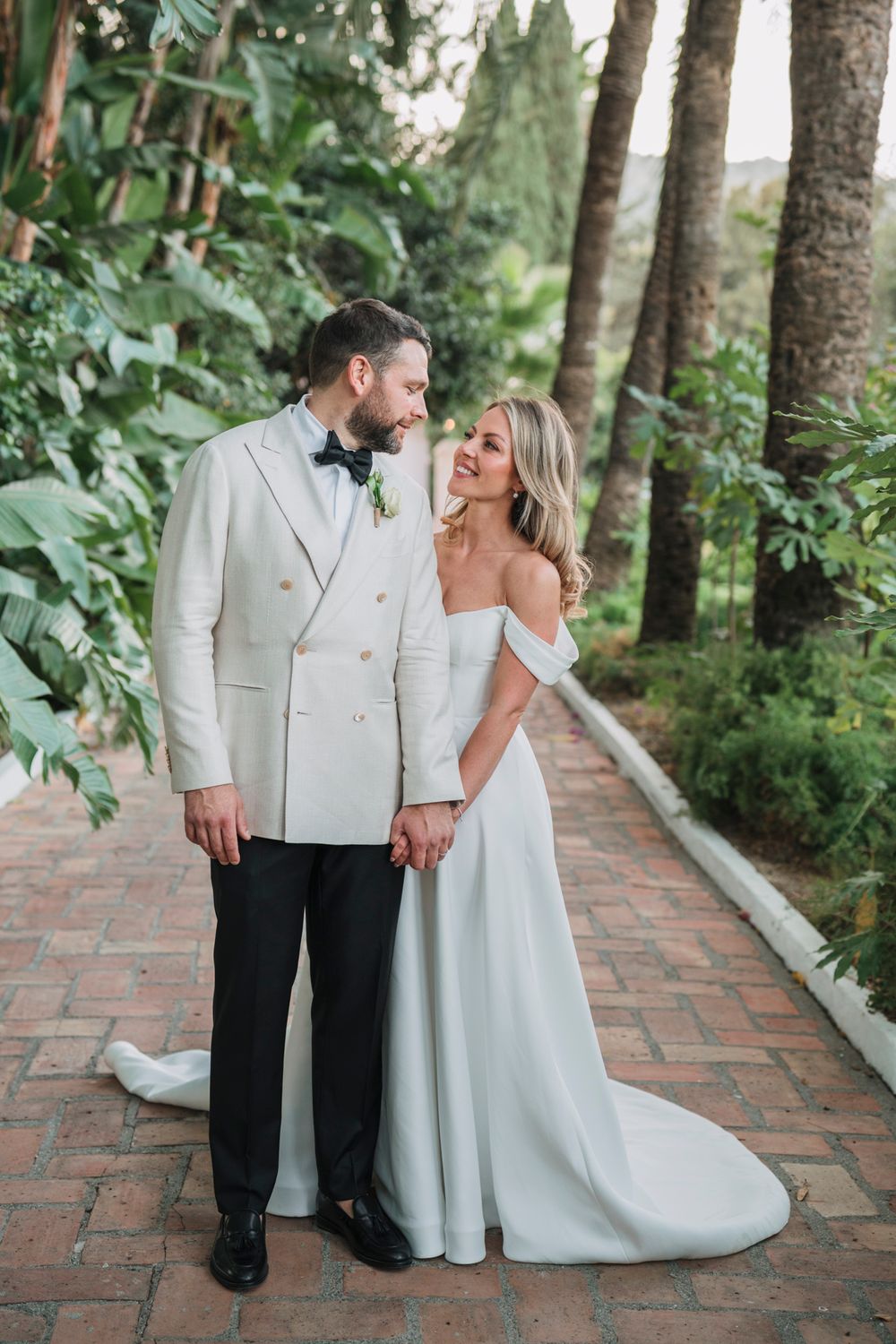 A romantic wedding portrait of a couple in white attire standing on a garden pathway.