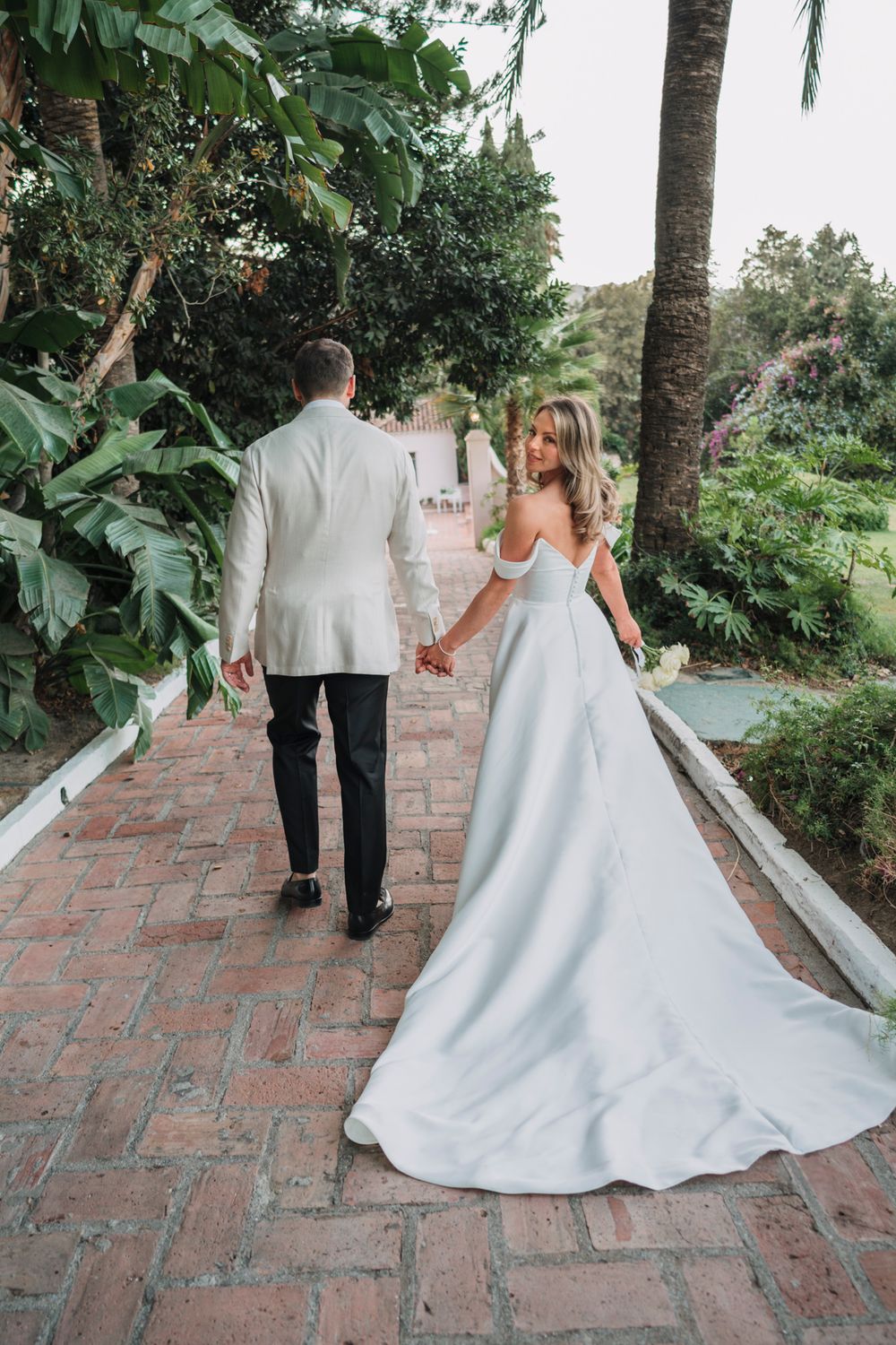 A couple walks hand in hand down a brick pathway surrounded by tropical gardens.
