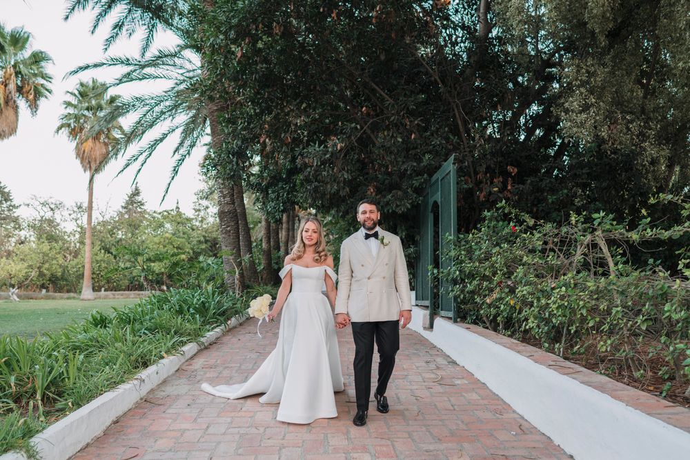A bride and groom walk along a palm-lined pathway in a garden setting.