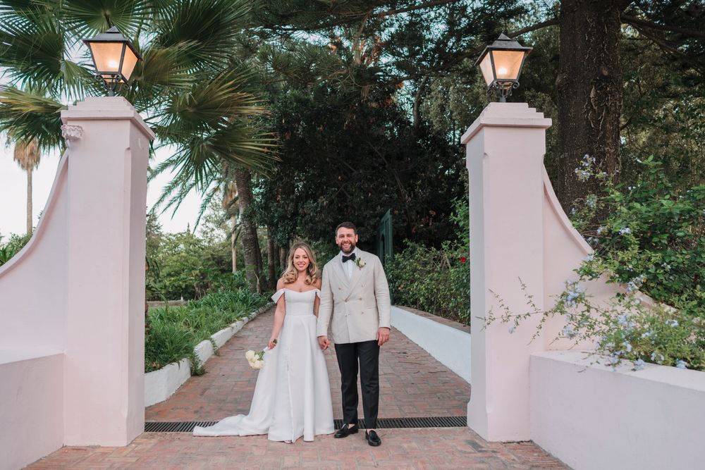 A wedding couple stands between two pink pillars with lanterns in a tropical garden setting.