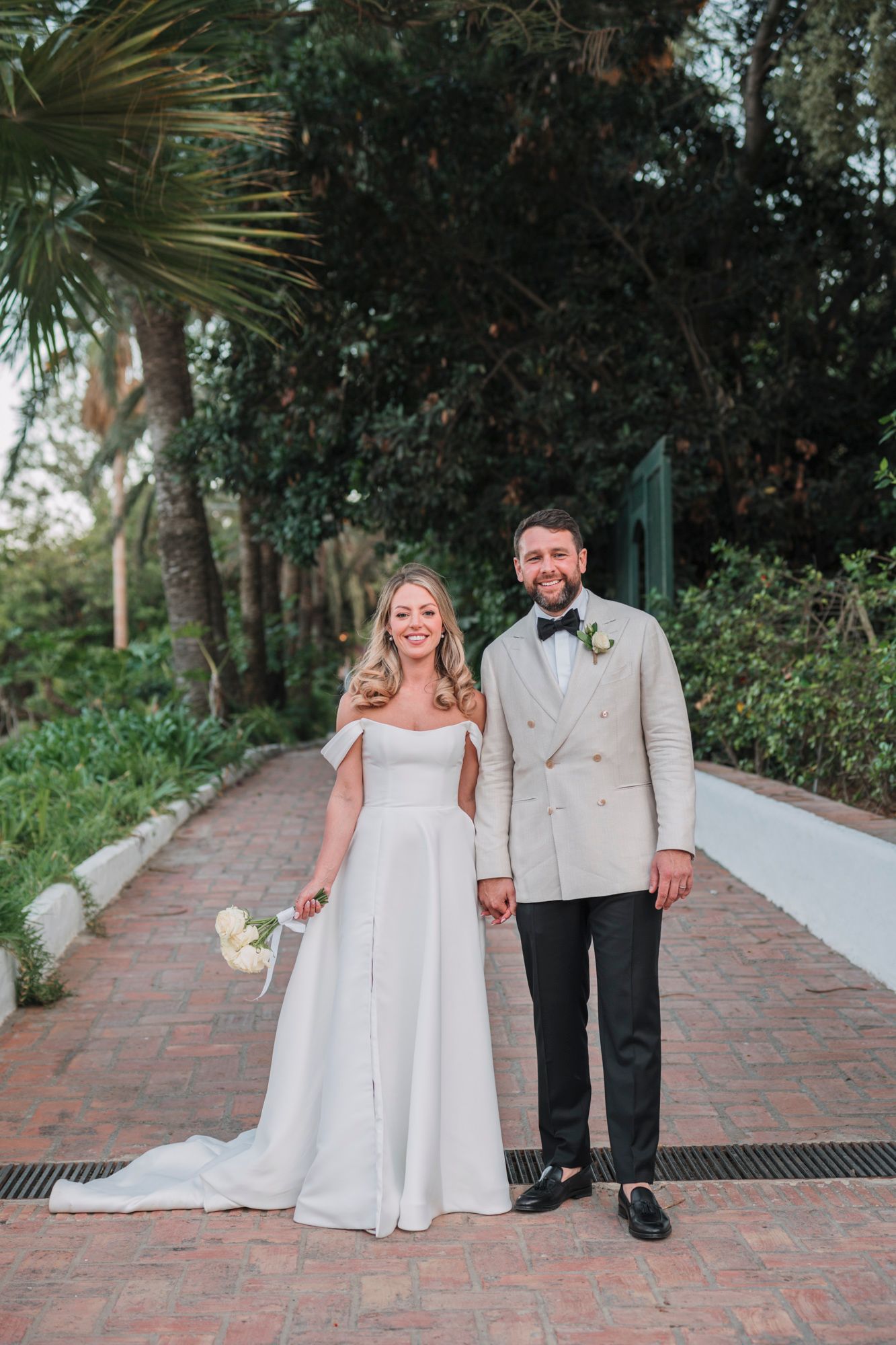 A happy couple poses on a brick pathway with palm trees and greenery in the background.