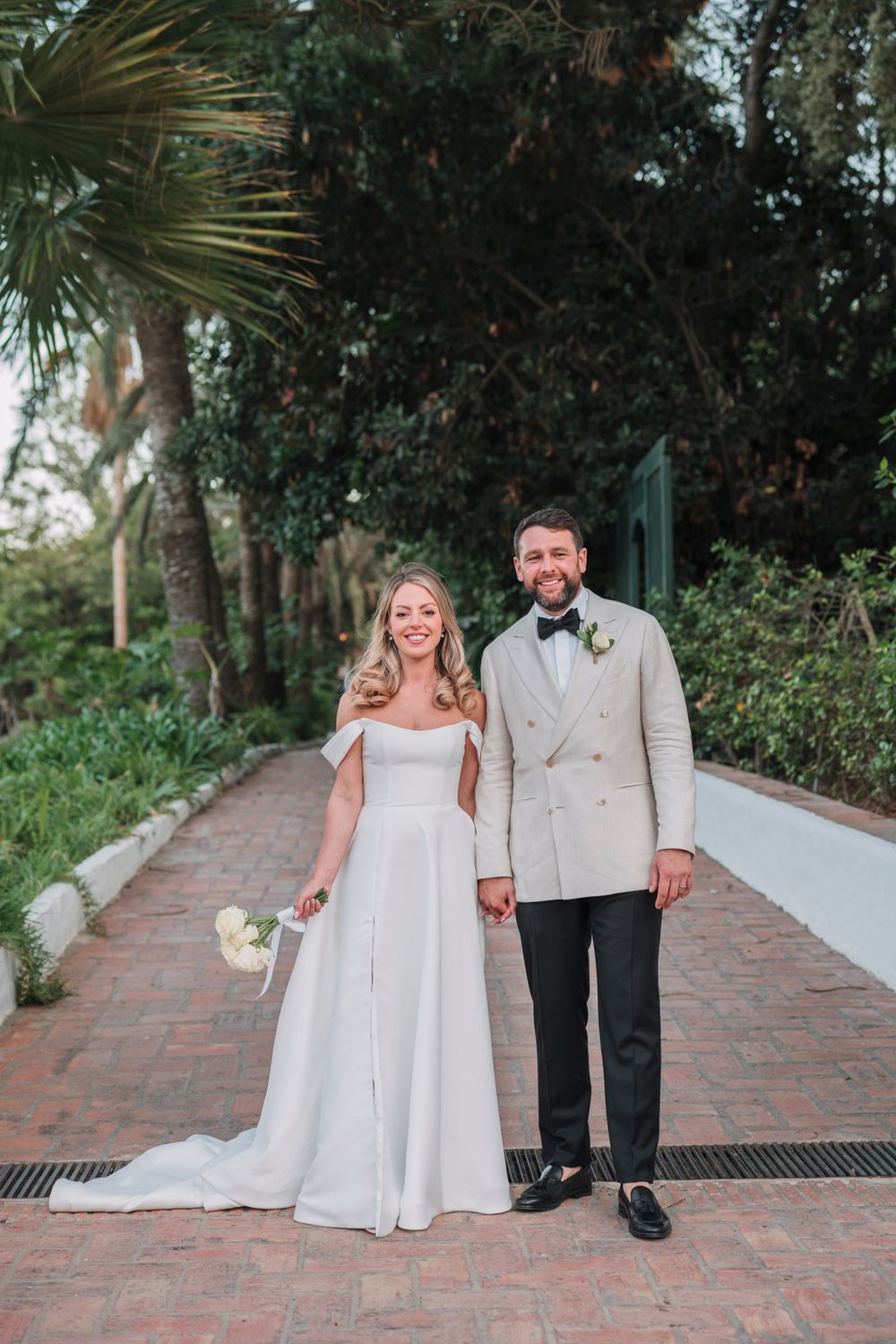 A happy couple poses on a brick pathway with palm trees and greenery in the background.